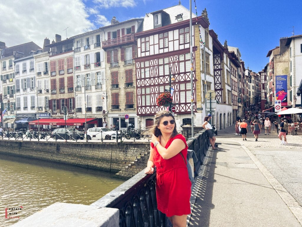 A woman in a bright red dress and sunglasses standing on a riverside promenade in Bayonne, France. Behind her are traditional Basque buildings with distinctive half-timbered facades and red shutters lining the Nive River. The historic city center features a mix of architectural styles with white buildings, colorful accents, and narrow streets filled with pedestrians under a bright blue sky.