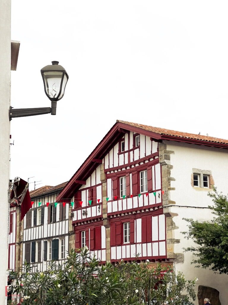 Traditional Basque architecture in a village in the French Basque Country, featuring distinctive white buildings with red and maroon half-timbering and shutters. The traditional Labourd-style houses display typical elements including exposed wooden beams, steep rooflines with terracotta tiles, and stone corner details, with small festive flags strung between buildings.
