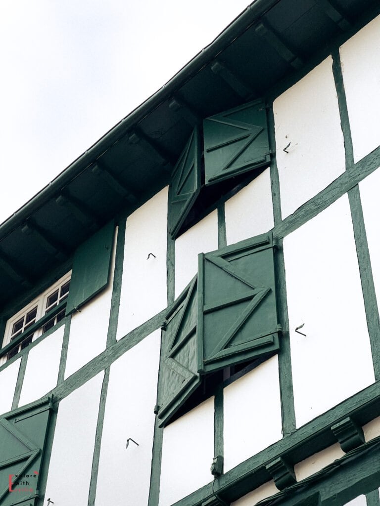 Close-up detail of traditional Basque architecture in Espelette village, featuring a white building with distinctive green half-timbering and matching green shutters. The characteristic colombage (timber framing) creates geometric patterns against the white plaster walls, with several shutters partially open, showcasing the distinctive craftsmanship and design elements typical of the French Basque Country building style.