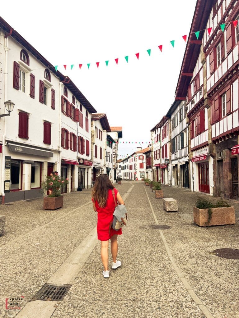 A woman in a bright red dress walking down a picturesque cobblestone street in Espelette village in the French Basque Country. The street is lined with traditional white buildings featuring distinctive red wooden shutters and half-timbered facades. Colorful Basque flag bunting (red, white, and green) hangs between buildings, and decorative planters line the pedestrian-friendly thoroughfare.