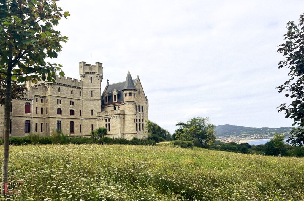 Abbadia Castle (Château d'Abbadia) near Hendaye in the French Basque Country, featuring neo-Gothic and neo-Medieval architecture with distinctive towers, crenellations, and stone masonry. The impressive castle stands on a hillside surrounded by a wildflower meadow, with distant views of the Bay of Txingudi and mountains visible on the horizon. This 19th-century landmark was designed by Eugène Viollet-le-Duc and built for explorer and scientist Antoine d'Abbadie.