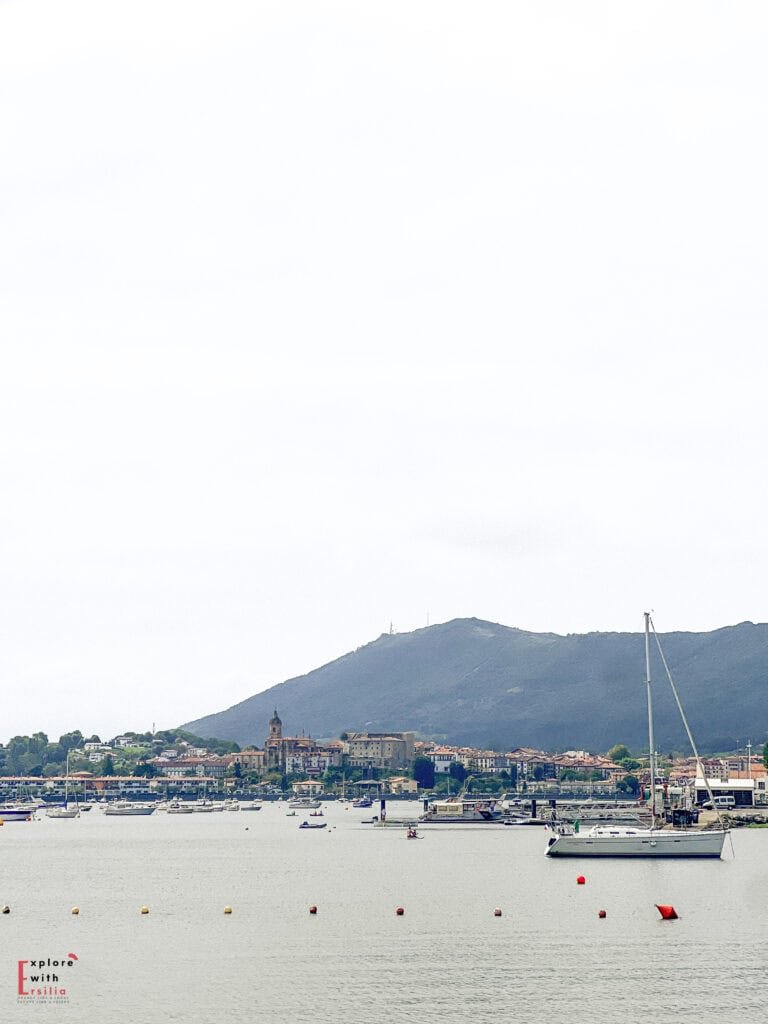 View across the Txingudi Bay from Hendaye, France, looking toward Hondarribia (Fuenterrabía) in Spain. The panorama shows sailboats moored in the calm harbor waters with the Spanish Basque town visible on the opposite shore. Traditional buildings with red-tiled roofs cluster around a church with a prominent bell tower, all nestled at the foot of Mount Jaizkibel. This scene captures the transboundary nature of the Basque Country where France meets Spain across the bay.