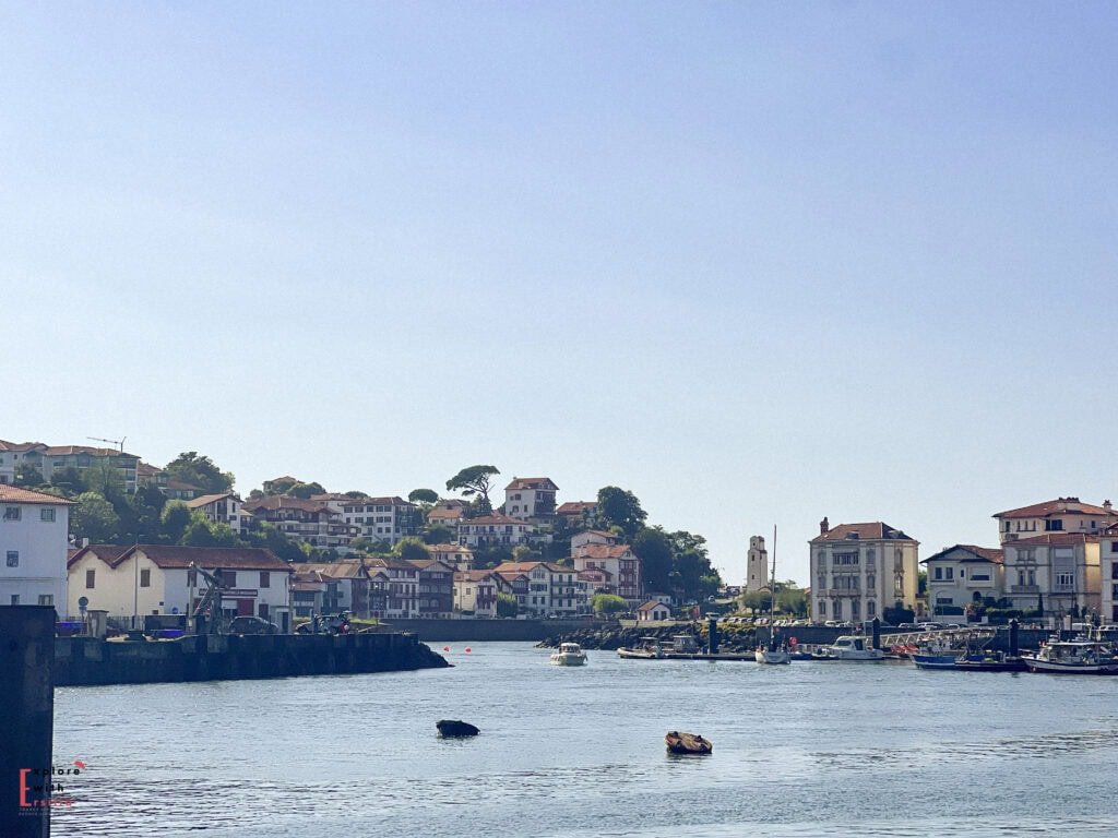 Panoramic harbor view of Saint-Jean-de-Luz, a picturesque fishing port and resort town in the French Basque Country. The image shows the town's characteristic white buildings with red roofs cascading down the hillside around the protected bay. Small fishing boats and pleasure craft are moored in the calm harbor waters, with the historic town center visible on the right featuring traditional Basque architecture, and a church tower (likely the Church of Saint John Baptist) rising among the buildings under a clear blue sky.