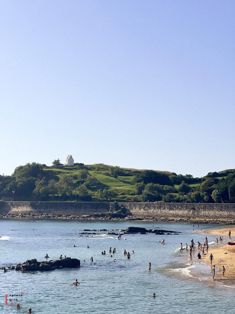 Beach scene in Saint-Jean-de-Luz with swimmers enjoying the sheltered bay waters, while Colline Sainte-Barbe rises in the background. This green hillside features a distinctive white chapel or lookout building at its summit. Stone seawalls protect the beach area, with rocky outcrops visible in the water where beachgoers are swimming. The scene captures the natural beauty of this French Basque coastal town under a clear blue sky.