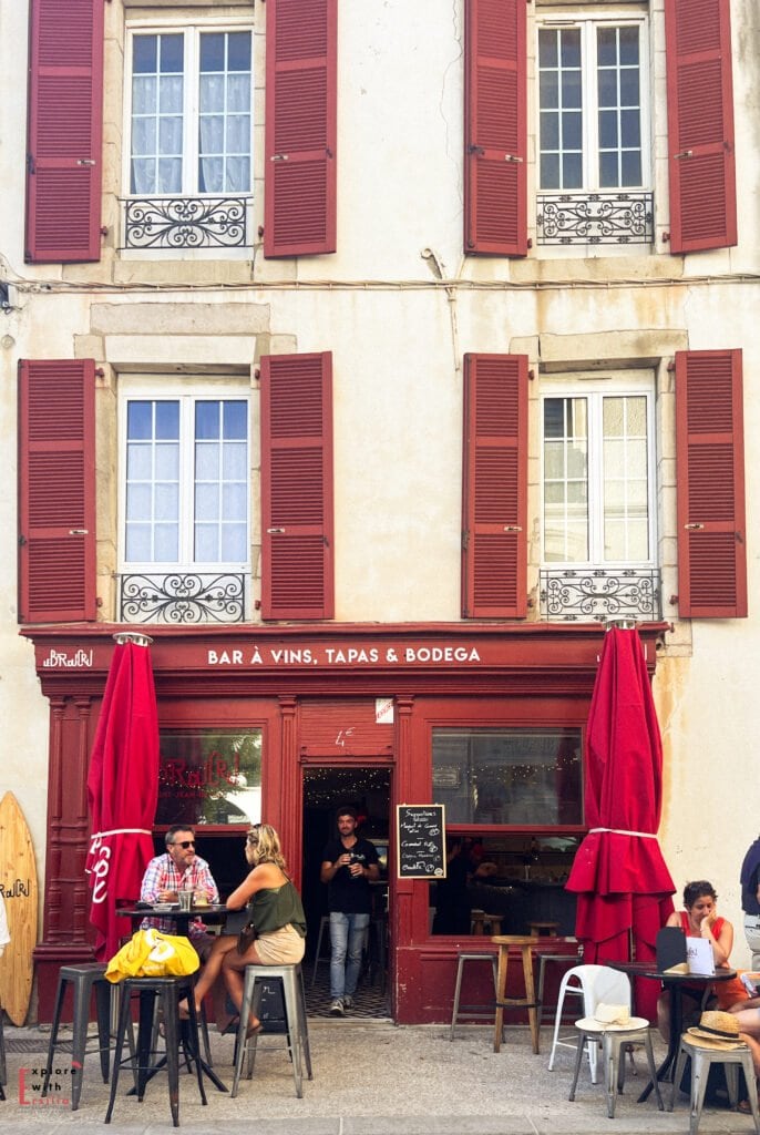 Charming wine bar and tapas restaurant in Saint-Jean-de-Luz with distinctive red facade and matching shutters on a traditional cream-colored building. The storefront reads "BAR À VINS, TAPAS & BODEGA" with outdoor seating featuring high tables, stools, and red umbrellas where patrons are enjoying drinks. Decorative wrought iron balconies adorn the upper floor windows, showcasing the blend of French and Spanish Basque architectural influences.