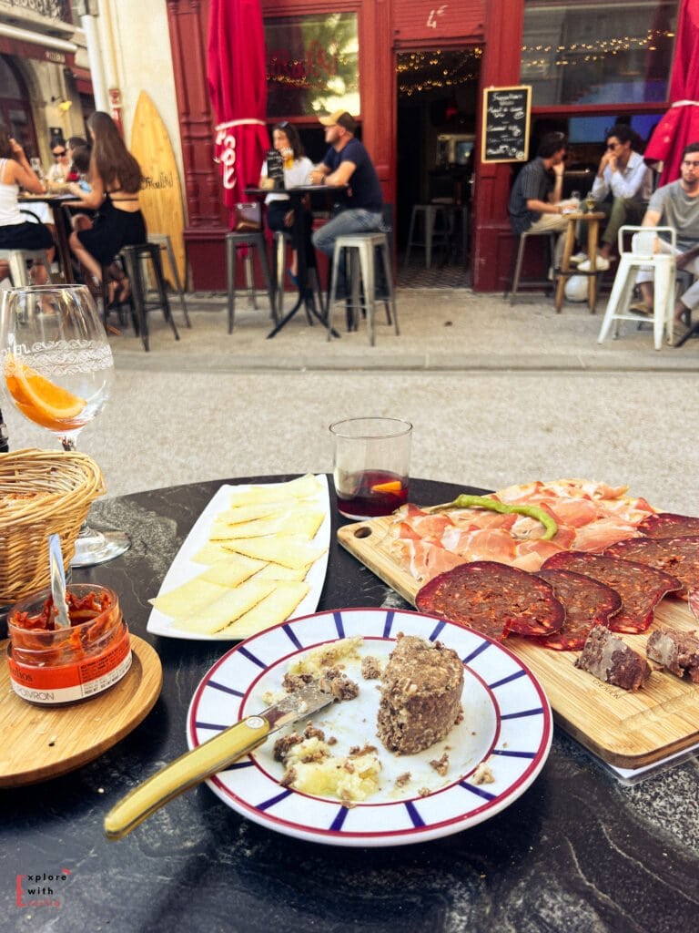 Table spread of Basque and Spanish tapas at an outdoor cafe in Saint-Jean-de-Luz, featuring a wooden charcuterie board with jamón ibérico (Spanish ham) and chorizo, a plate of Manchego cheese, pâté or rillete on a decorative plate, and a glass of sangria with orange slice. In the background, the same red-fronted wine bar terrace is visible with customers enjoying drinks at high tables.
