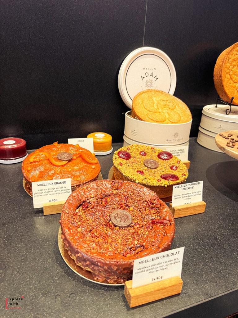 Display of gourmet cakes and pastries from Maison Adam in the French Basque Country. Three elegant round cakes are prominently featured: a chocolate "Moelleux Chocolat" (19.90€), an orange "Moelleux Orange" (19.90€), and a pistachio-raspberry "Moelleux Framboise Pistache". In the background are traditional Basque cookies (gâteau basque) in decorative round tins with the Maison Adam logo, showcasing this renowned patisserie's regional specialties.