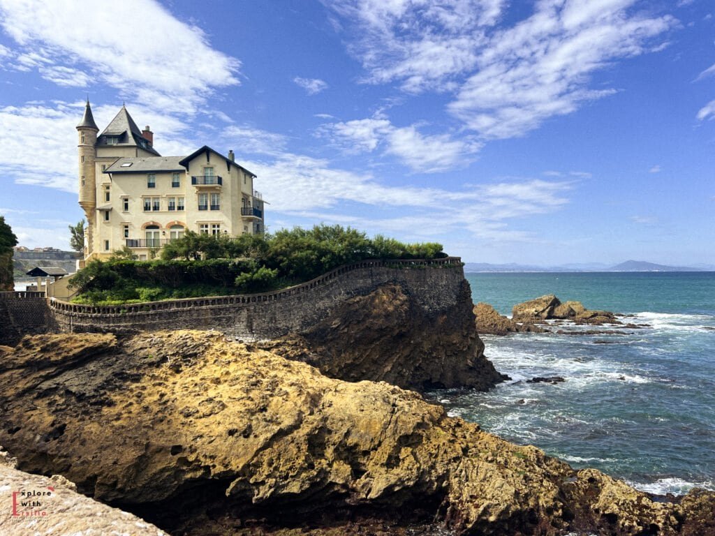 Historic villa with a turret perched dramatically on a rocky cliff overlooking the Atlantic Ocean in Biarritz, France. The elegant cream-colored building sits behind a stone wall along the rugged coastline, with waves crashing against the rocks below. The scene is set against a brilliant blue sky with scattered white clouds, and the Bay of Biscay stretches to the horizon with distant mountains visible.