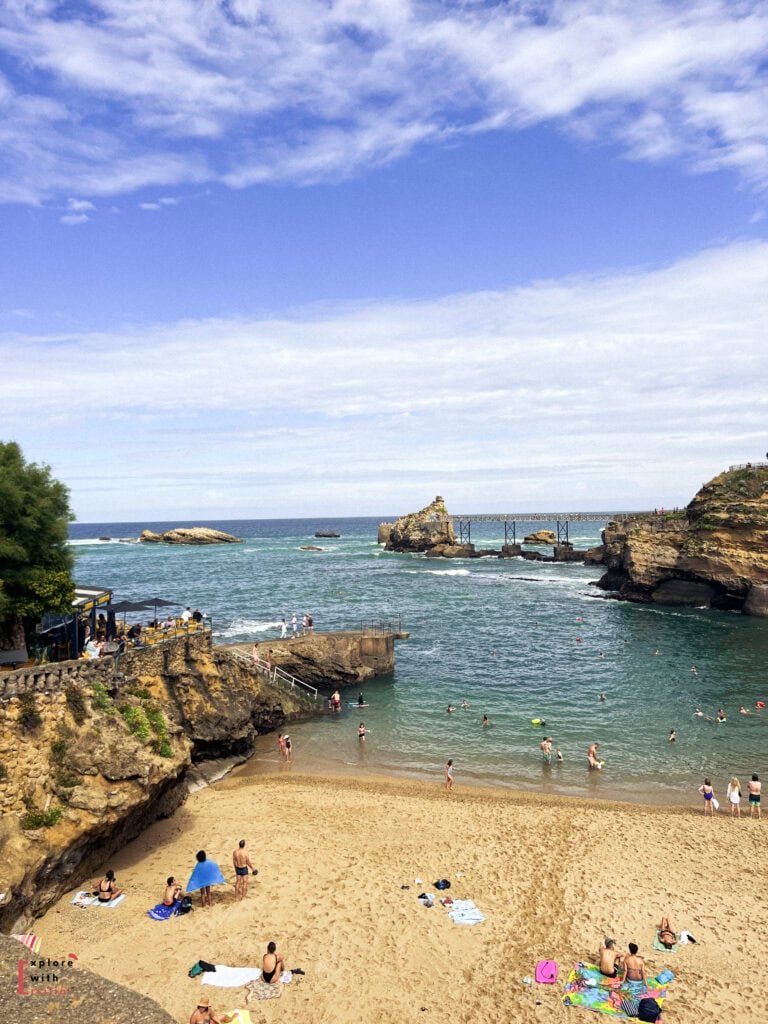 Beautiful view of Plage du Port Vieux in Biarritz, a small sheltered beach cove surrounded by rocky cliffs. The sandy beach is dotted with sunbathers, while swimmers enjoy the turquoise waters of this natural harbor. A distinctive footbridge connects rock formations in the distance, and swimming platforms are built into the rocky coastline. The scene is set against a bright blue sky with scattered clouds over the Atlantic Ocean.