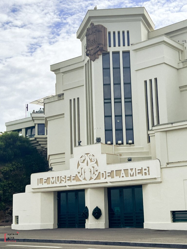 The striking Art Deco facade of Le Musée de la Mer (Sea Museum) in Biarritz, featuring a white exterior with vertical blue glass windows and geometric architectural elements. The building's modernist design includes prominent vertical lines, a stepped entrance with the museum name displayed prominently, and decorative maritime motifs. This landmark cultural institution is set against a cloudy blue sky in the French Basque coastal resort town.