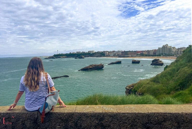 A person in a striped shirt sits on a stone wall overlooking the panoramic coastal view from Atalaye Plateau in Biarritz. The scene captures the city's dramatic Atlantic coastline with rocky outcrops, sandy beaches, and urban development stretching along the curved bay. The Biarritz lighthouse is visible on a distant headland, and the town's elegant buildings line the shore under a dramatic sky with blue and white clouds.