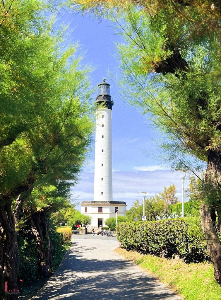 The tall white Biarritz Lighthouse (Phare de Biarritz) standing prominently against a bright blue sky, framed by lush green pine trees. A path leads to the cylindrical tower with its black lantern room and distinctive black spiral band. The base building houses visitor facilities, with landscaped grounds surrounding this iconic maritime landmark on the French Basque coast.