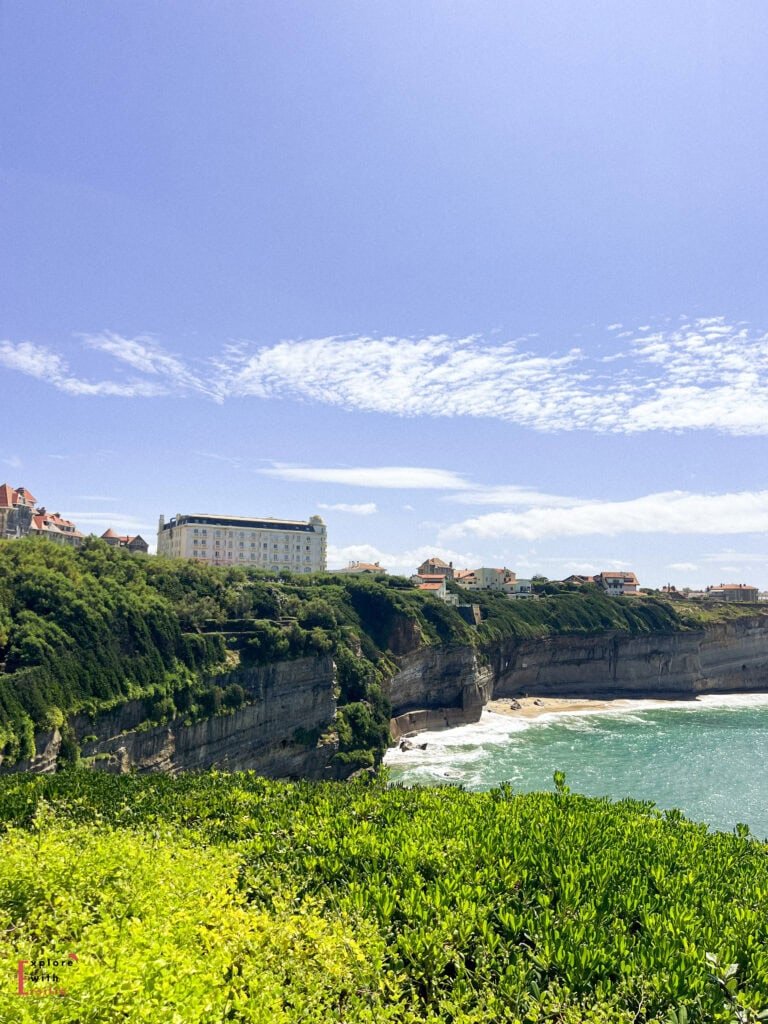 Dramatic coastal view of Biarritz showing luxury Belle Époque buildings perched atop steep limestone cliffs overlooking a small beach cove. The scene captures the characteristic landscape of this elegant resort town, with verdant vegetation covering the cliff faces, turquoise Atlantic waters below, and a grand white hotel or apartment building prominently positioned along the cliff edge under a bright blue sky with wispy clouds.