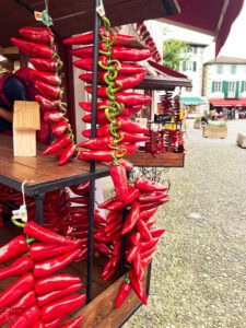 Vibrant strings of red Espelette peppers hanging from a market stall in a French Basque village. Multiple garlands of the glossy peppers are displayed at a wooden market stand, with traditional Basque buildings visible in the background and a stone plaza in the foreground.