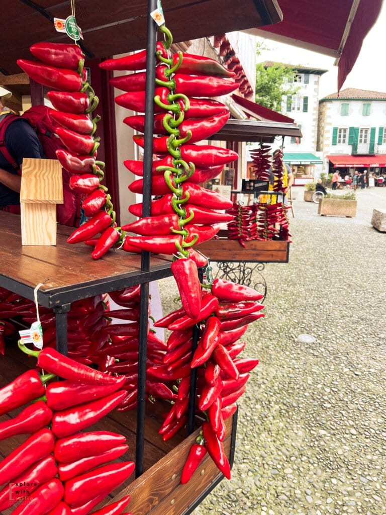Vibrant strings of red Espelette peppers hanging from a market stall in a French Basque village. Multiple garlands of the glossy peppers are displayed at a wooden market stand, with traditional Basque buildings visible in the background and a stone plaza in the foreground.