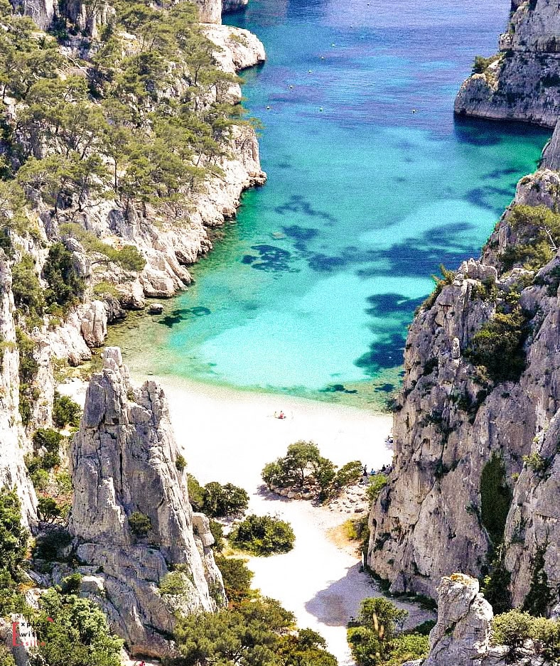 Breathtaking aerial view of a calanque near Marseille, with towering limestone cliffs enclosing a secluded cove of turquoise and deep blue water. A narrow white-sand beach sits at the base, dotted with a few visitors, and surrounded by lush Mediterranean pines and rugged rock formations.