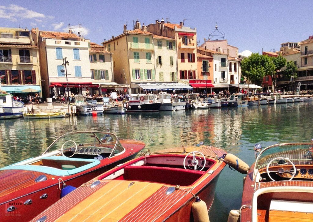 Colorful harbor scene in Cassis, France, with vintage wooden boats docked in calm turquoise water and pastel buildings lining the bustling waterfront. Cafés and restaurants with outdoor seating fill the promenade, creating a lively Mediterranean atmosphere under a clear blue sky.