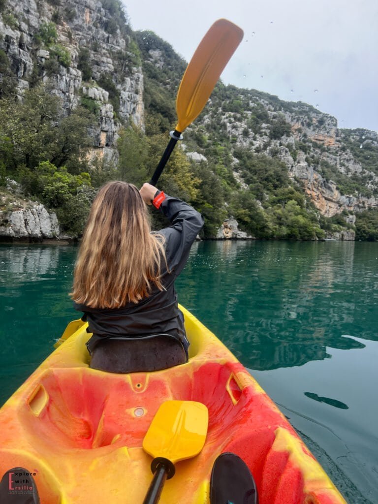 Person kayaking through the emerald waters of the Gorges du Verdon, seen from behind in a bright red and yellow kayak, paddling between steep, rocky cliffs covered in lush greenery. The scene captures the adventurous and serene spirit of exploring this dramatic natural canyon in Provence.