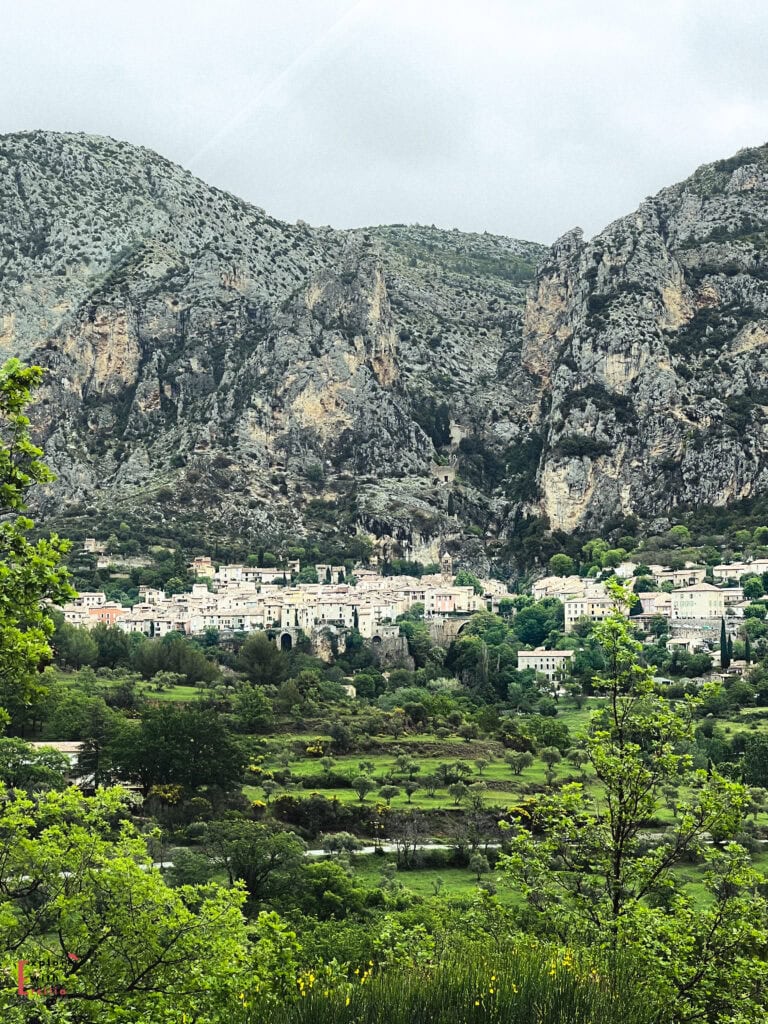 Scenic view of Moustiers-Sainte-Marie, a picturesque village in Provence nestled at the base of dramatic limestone cliffs, with pastel-hued buildings clustered against the rugged rock face. Lush green fields and trees fill the foreground under an overcast sky, highlighting the village's stunning natural setting.