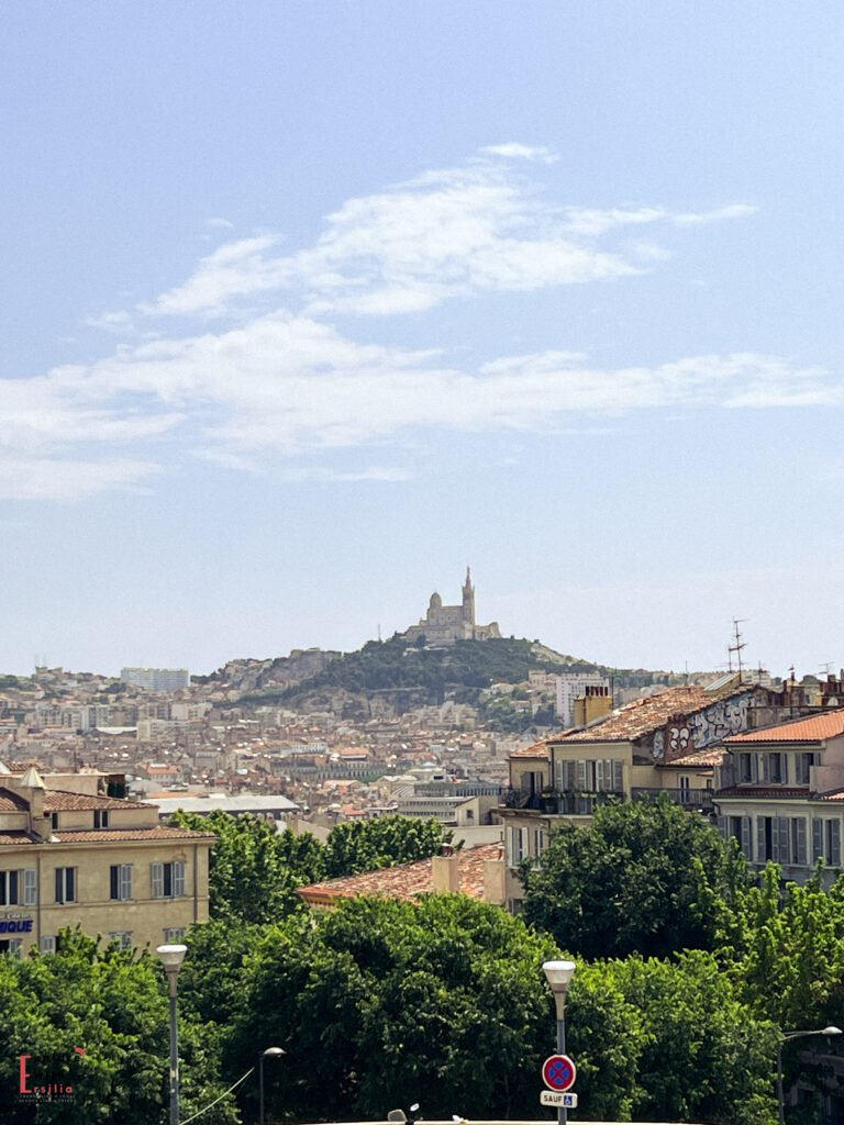 Panoramic view of Marseille with the iconic Notre-Dame de la Garde basilica perched atop a hill in the distance, overlooking the city’s dense rooftops and urban landscape. The foreground includes classic Mediterranean buildings with terracotta tiles and leafy green trees under a bright blue sky.