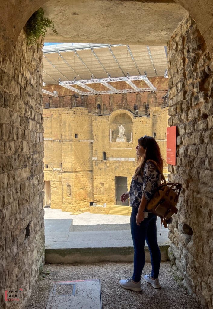 A woman with a backpack stands in a stone passageway, looking out at the stage wall of the Roman Theatre of Orange in France. The grand backdrop features niches, arches, and a statue of Emperor Augustus, partially covered by a modern metal roof structure above the ancient ruins.