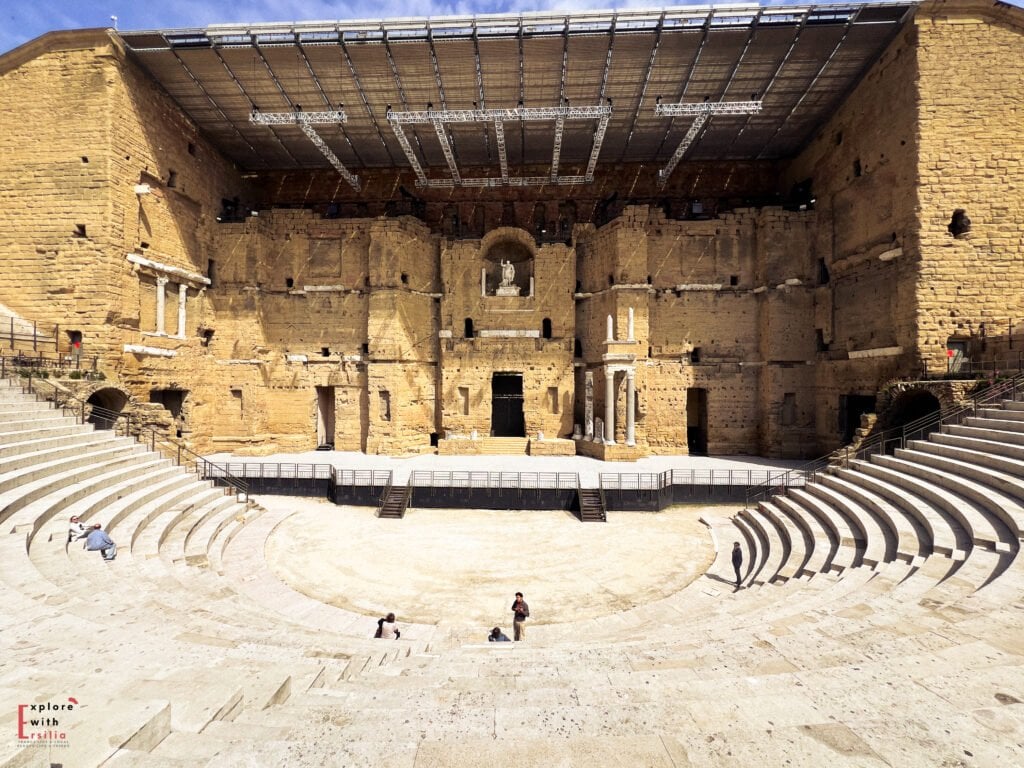 Wide-angle view of the Roman Theatre of Orange in Provence, showcasing its remarkably preserved stage wall, stone seating tiers, and modern overhead structure. A few visitors are scattered around the ancient amphitheater, highlighting its grand scale and historic ambiance.