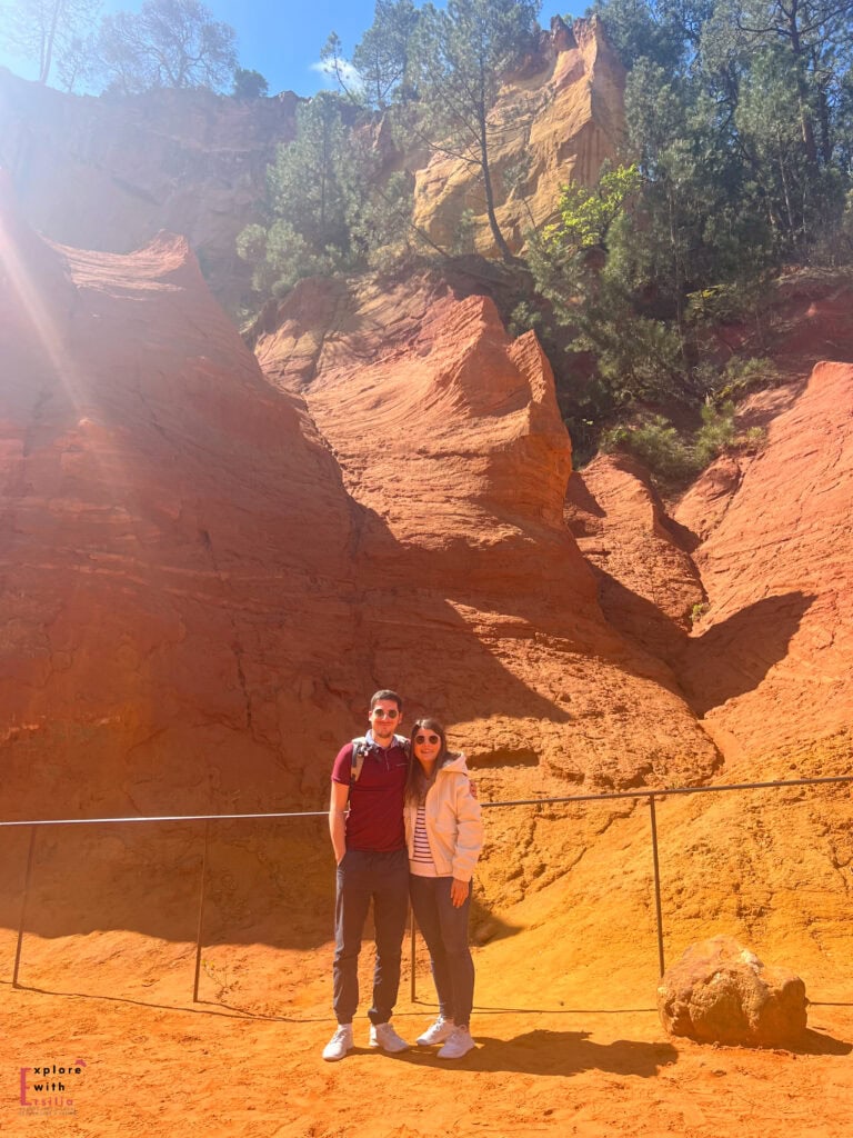 A smiling couple stands in front of vibrant red and orange ochre cliffs at Sentier des Ocres, with sunlight casting a soft glow over the surreal landscape and trees perched on the cliffs above.