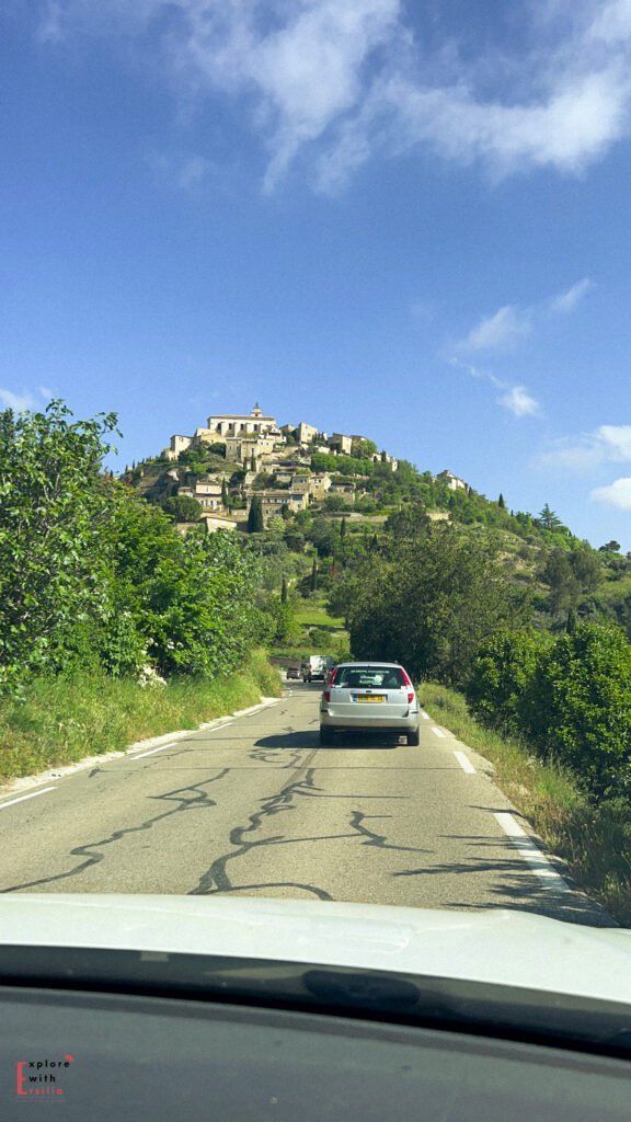 A scenic drive toward the hilltop village of Gordes in Provence, France, with a car traveling a narrow country road lined with greenery and the iconic stone buildings of Gordes rising above on a lush hillside under a bright blue sky.
