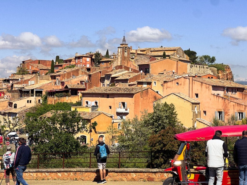 A vibrant hilltop village in Roussillon, Provence, with terracotta and ochre-colored houses stacked closely together, viewed under a blue sky as tourists admire the scene and a tuk-tuk waits nearby.
