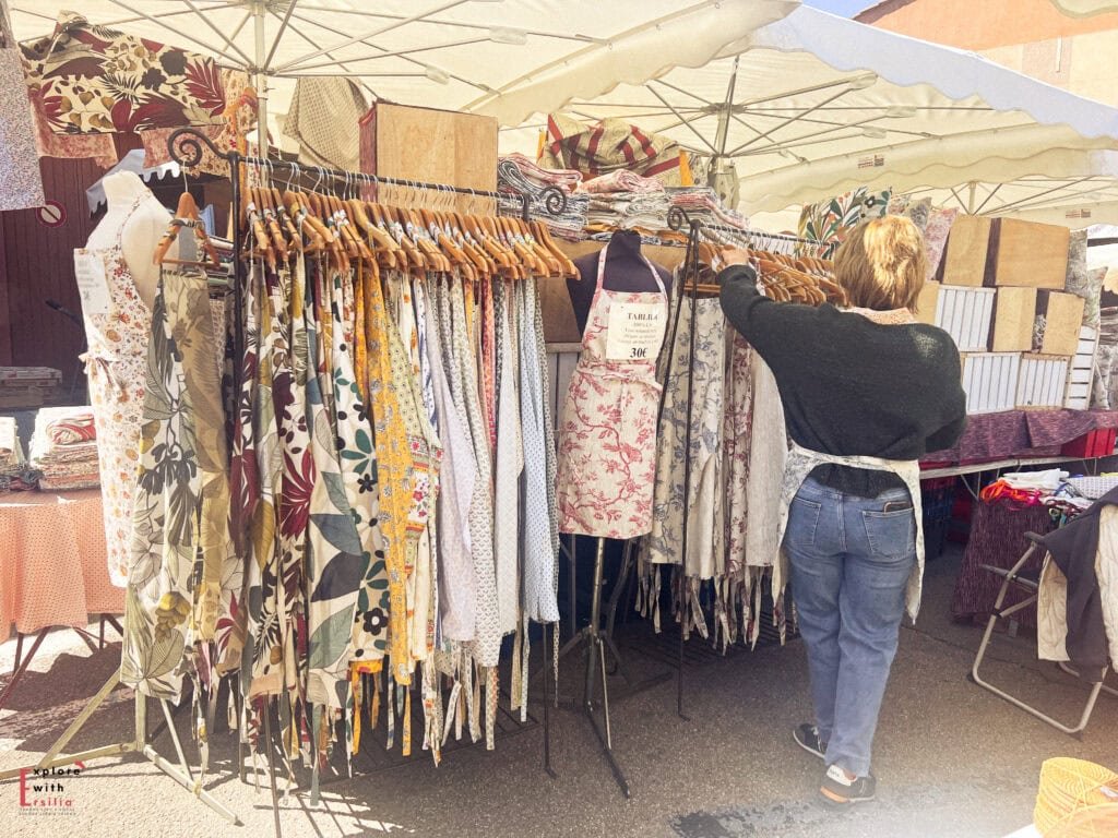 A woman browses colorful printed aprons at a Provence market stall, surrounded by hanging racks filled with floral and patterned textiles under a white canopy.