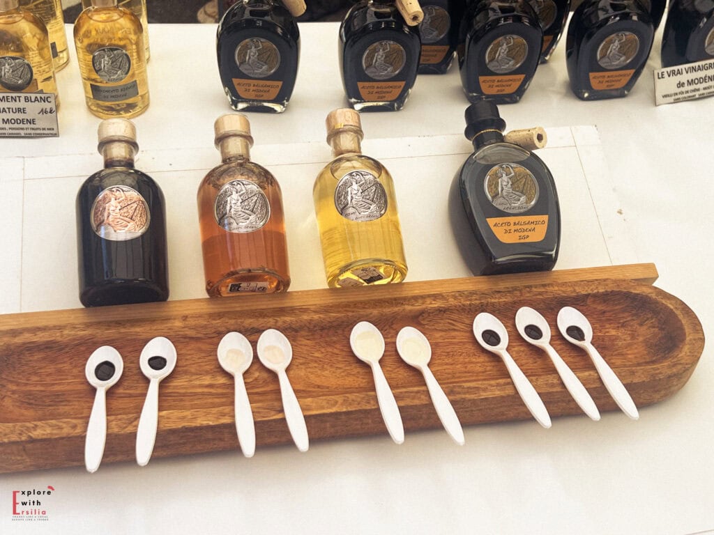A tasting setup for Aceto Balsamico di Modena at a market, featuring a wooden tray of sample spoons filled with light and dark vinegars, and elegantly labeled bottles displayed behind.
