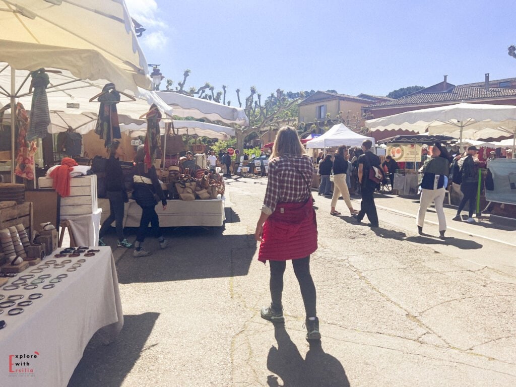 Shoppers explore a bustling outdoor market in Provence, lined with white canopy tents and stalls selling handmade goods, scarves, bags, and local treats under bright sunlight.