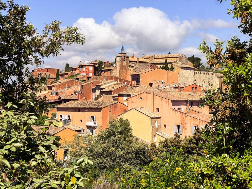 A panoramic view of the warm-toned, ochre-washed village of Roussillon in Provence, nestled among green trees with blue skies and soft clouds above.