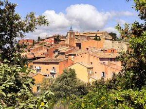 A panoramic view of the warm-toned, ochre-washed village of Roussillon in Provence, nestled among green trees with blue skies and soft clouds above.