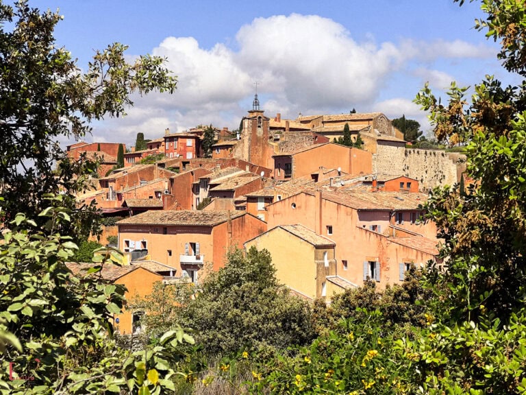 A panoramic view of the warm-toned, ochre-washed village of Roussillon in Provence, nestled among green trees with blue skies and soft clouds above.