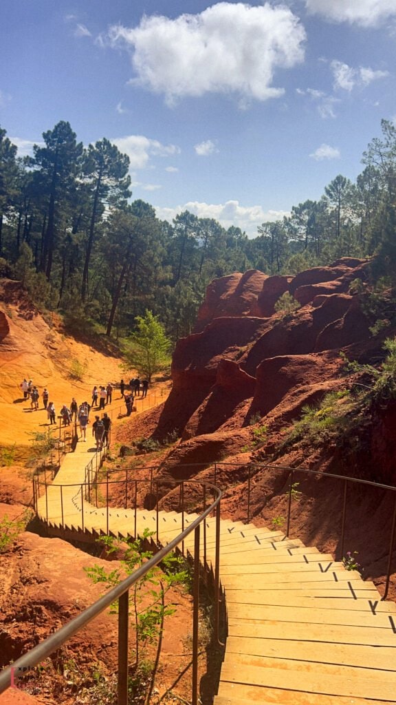 A wooden stairway descends through striking red ochre formations and sandy terrain, with groups of visitors exploring the colorful landscape surrounded by dense pine forest.