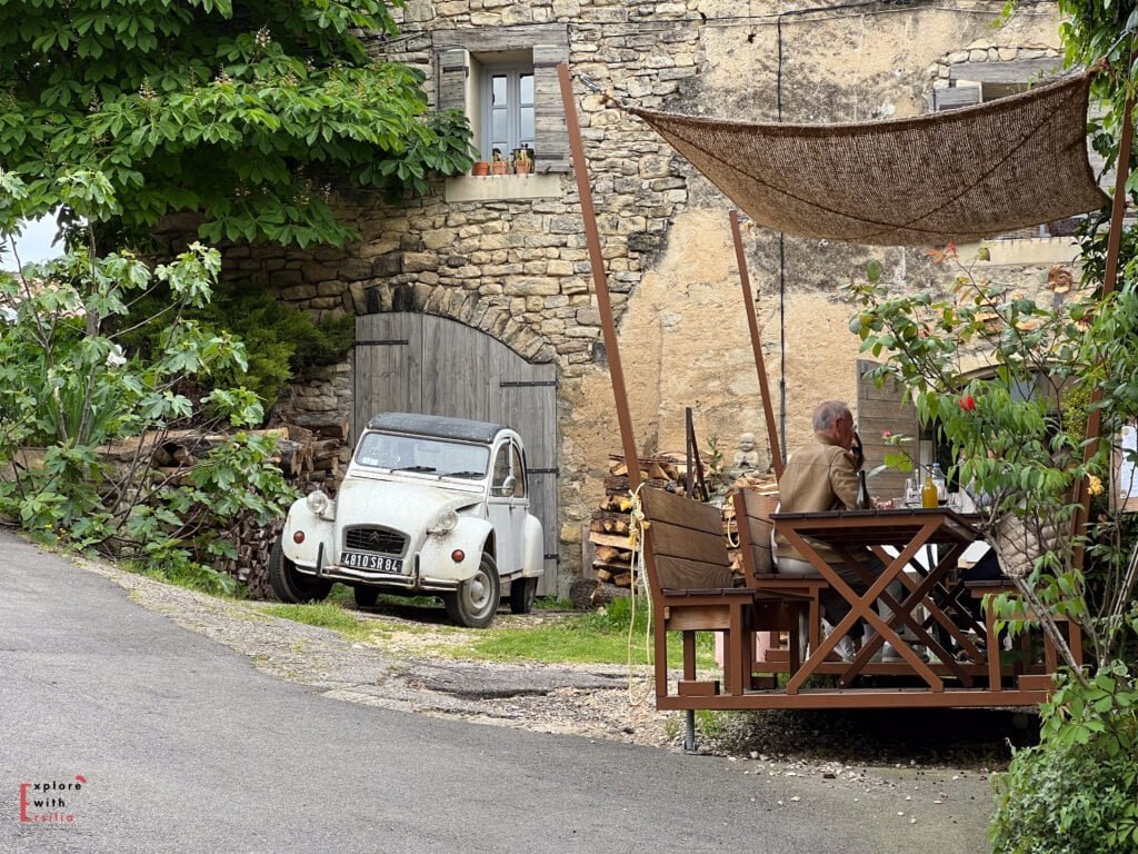 A vintage Citroën 2CV parked near a rustic stone house in Saignon village, with a man dining under a woven sunshade at a wooden patio—illustrating the timeless appeal of this hidden gem in Provence.
