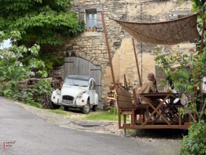 A vintage Citroën 2CV parked near a rustic stone house in Saignon village, with a man dining under a woven sunshade at a wooden patio—illustrating the timeless appeal of this hidden gem in Provence.