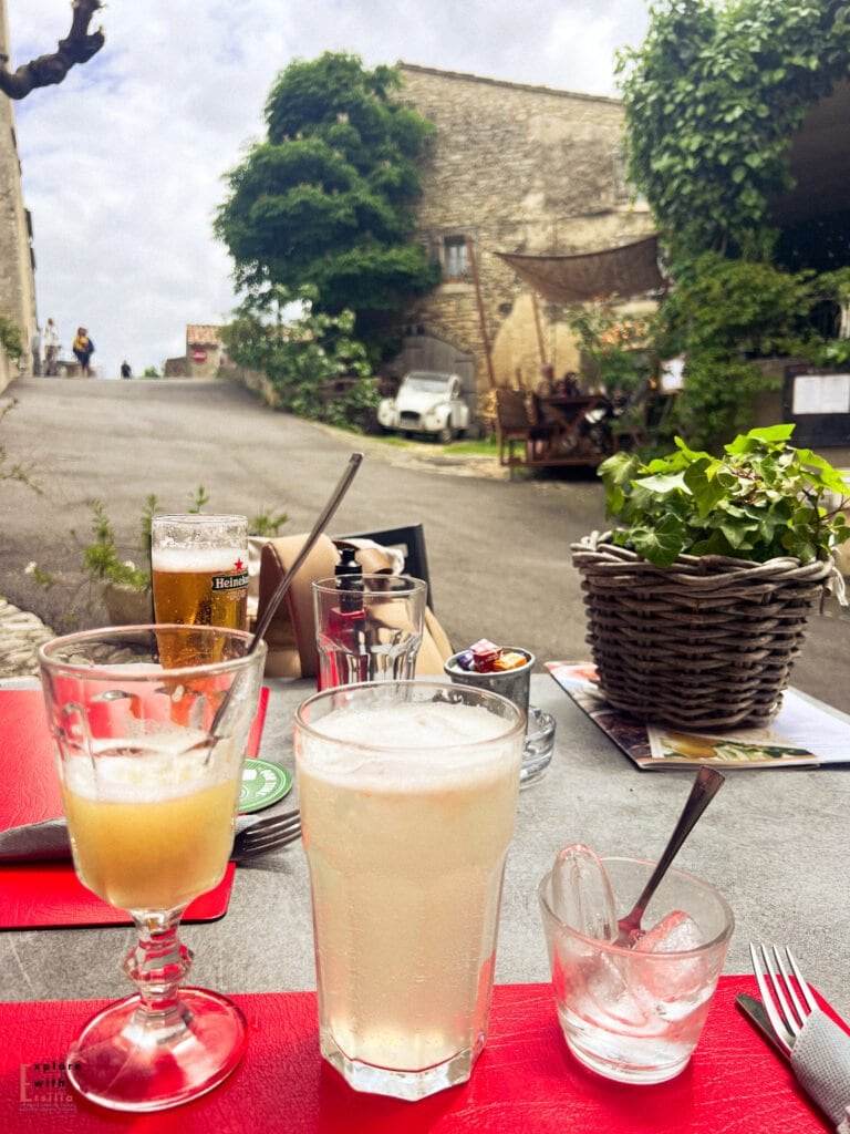 Outdoor café table in Saignon village set with refreshing drinks, including lemonade and beer, against a backdrop of stone buildings and winding roads—capturing the charm of this hidden gem in Provence.