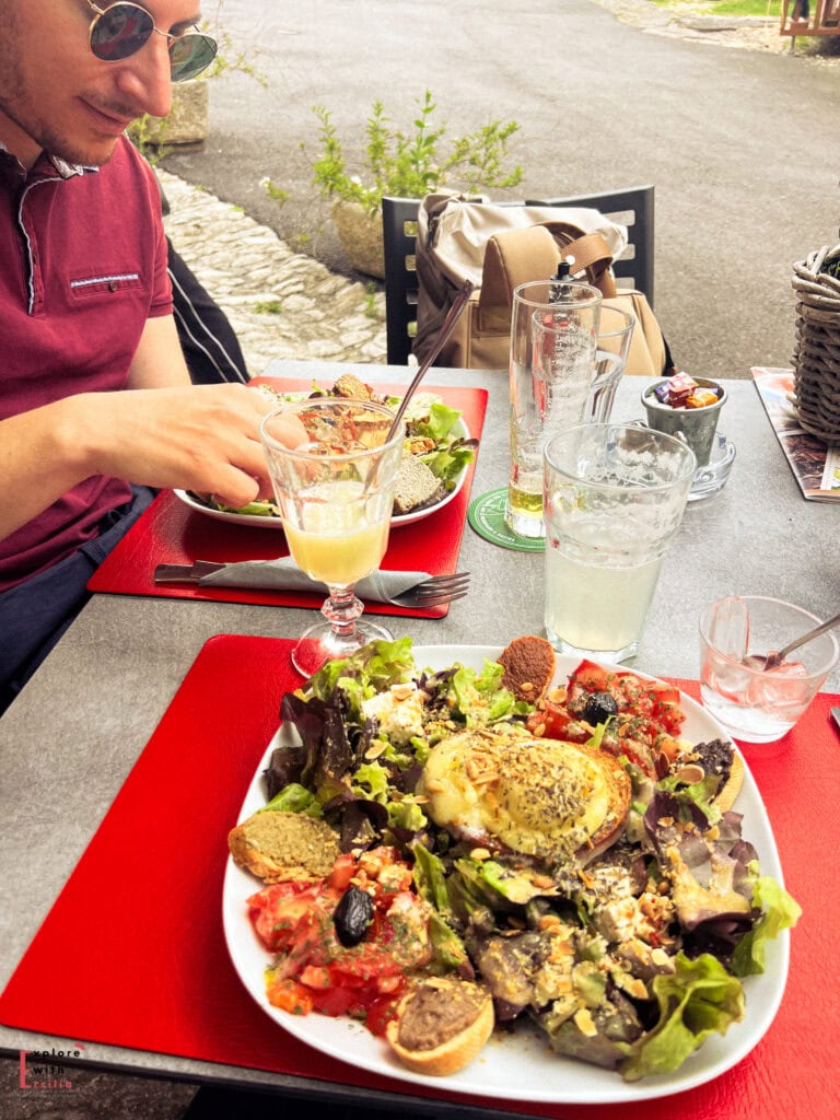 Fresh Provençal salad topped with baked goat cheese, olives, tomatoes, tapenade, and herbs, served al fresco on a red placemat in Saignon village, a hidden gem in Provence. A man in sunglasses enjoys his meal, with a rustic cobblestone street visible in the background.