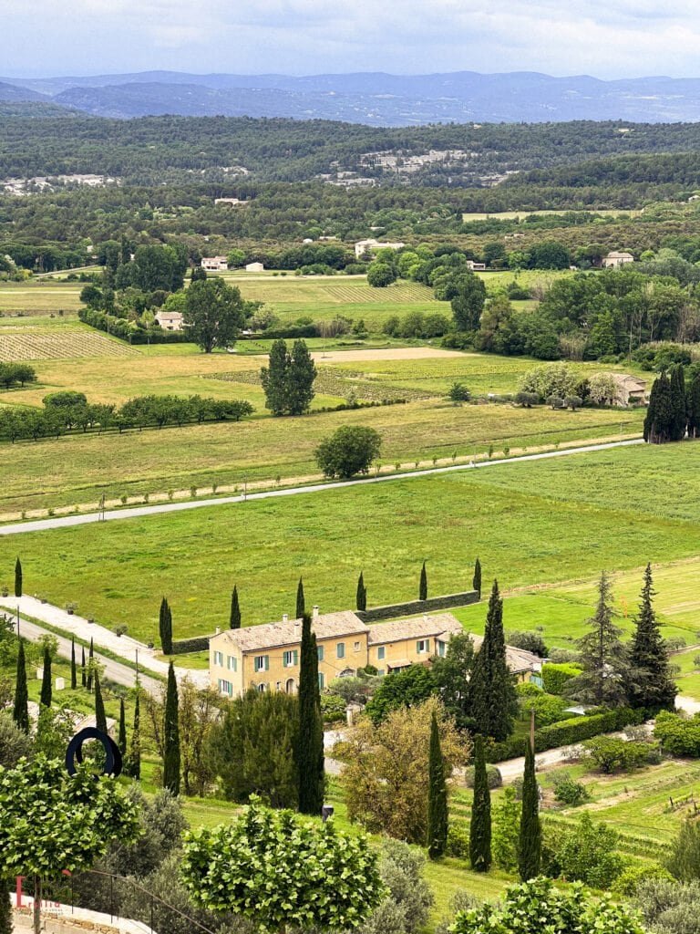 Scenic view of the countryside during a Provence village loop roadtrip, featuring rolling green fields, tall cypress trees, and a charming stone farmhouse nestled among vineyards and winding rural roads, with distant hilltop villages visible in the background.