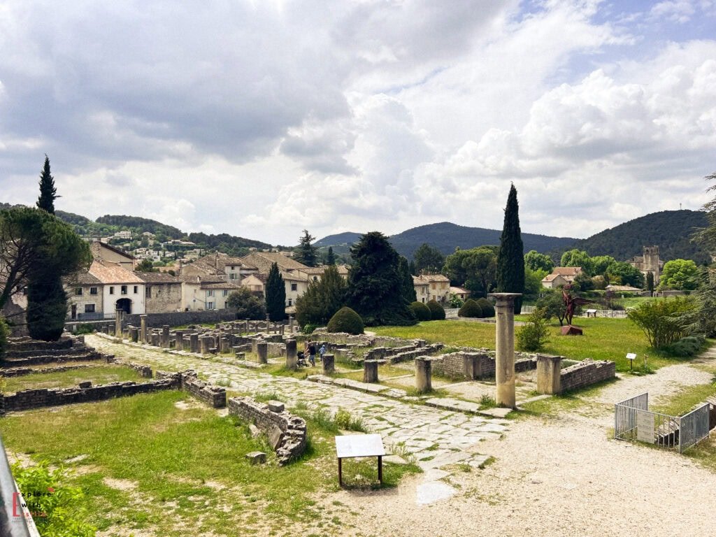View of Roman ruins in Vaison-la-Romaine, France, featuring a row of standing stone columns and remnants of ancient walls against a backdrop of a quaint village with terracotta rooftops. Rolling hills and cypress trees frame the scene under a dramatic sky filled with thick, textured clouds.