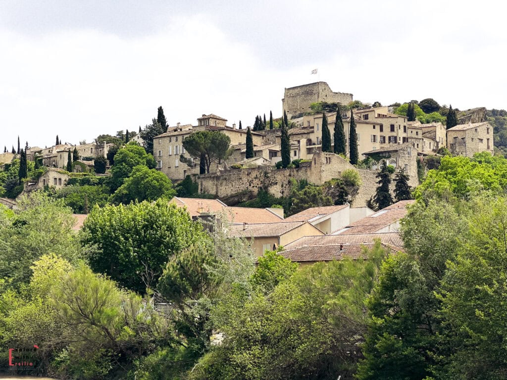 Panoramic view of the upper town of Vaison-la-Romaine in Provence, showcasing stone buildings with terracotta roofs nestled among tall cypress trees and lush greenery. A medieval castle ruin with a flag flying atop crowns the hill, adding to the village's historic charm.