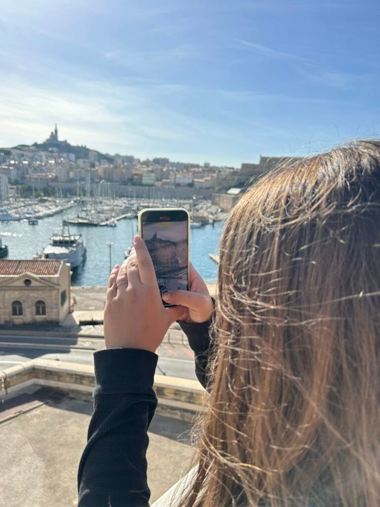 A traveler takes a photo of Marseille’s Old Port with a smartphone, capturing boats in the harbor and the iconic Notre-Dame de la Garde basilica on the hill. Perfect moment to highlight why using the best apps for your French travel can help you find scenic spots like this and document your journey effortlessly.