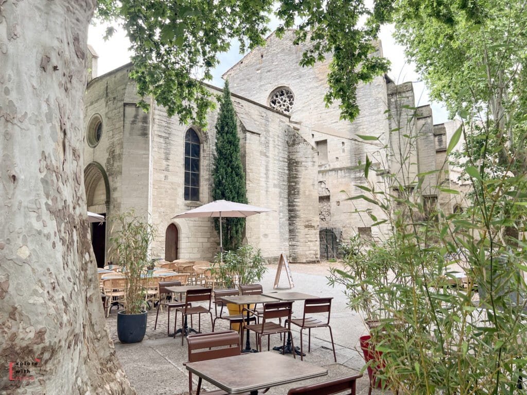 An outdoor café with tables and umbrellas sits beneath shady trees next to a historic stone church in Avignon, France. The peaceful setting combines medieval architecture with a relaxed, inviting atmosphere for dining al fresco.