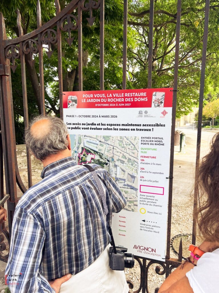 Two visitors read an information board about restoration work at the Jardin du Rocher des Doms in Avignon, France. The sign details construction phases from October 2024 to June 2027, with a map showing accessible areas and opening hours during the renovations.