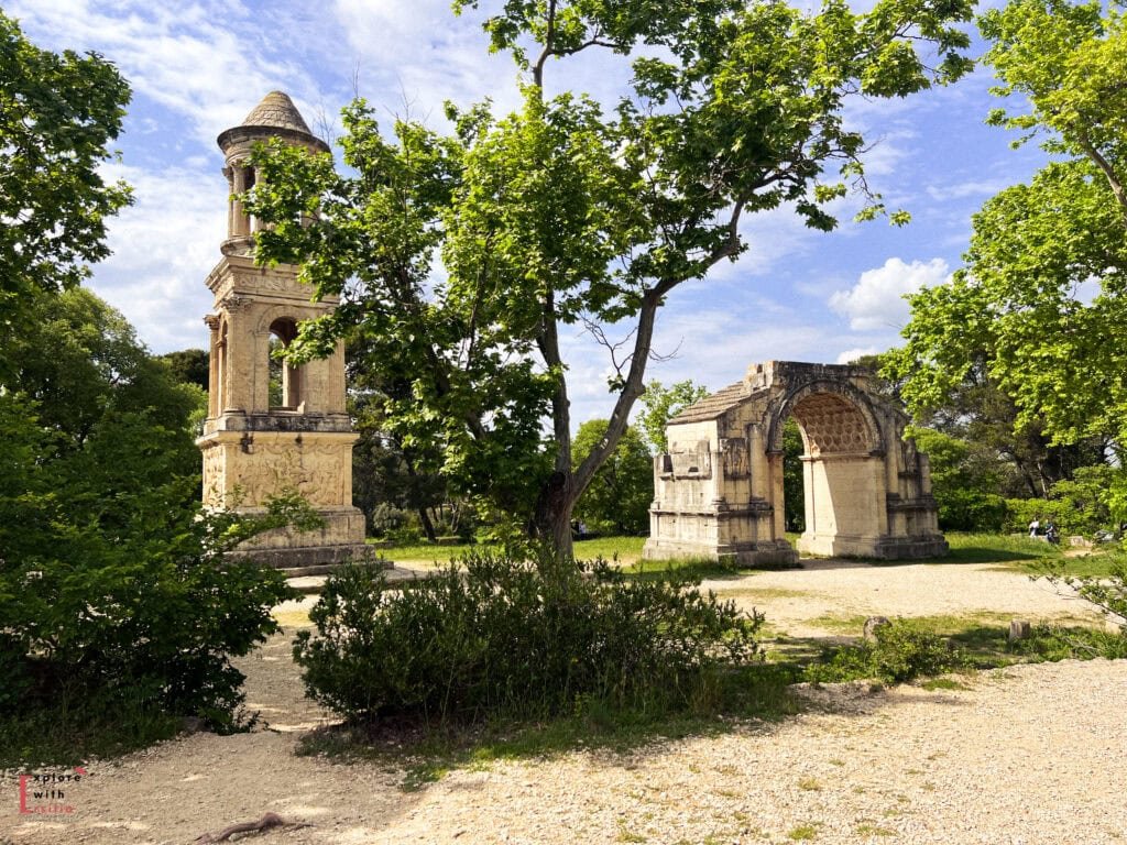 Two well-preserved Roman monuments—an ornate mausoleum and a triumphal arch—stand side by side among lush trees and gravel pathways in Glanum, Saint-Rémy-de-Provence. The mausoleum features intricate carvings and a cylindrical top, while the arch has decorative stone reliefs and a large central opening beneath a partially intact vaulted ceiling.
