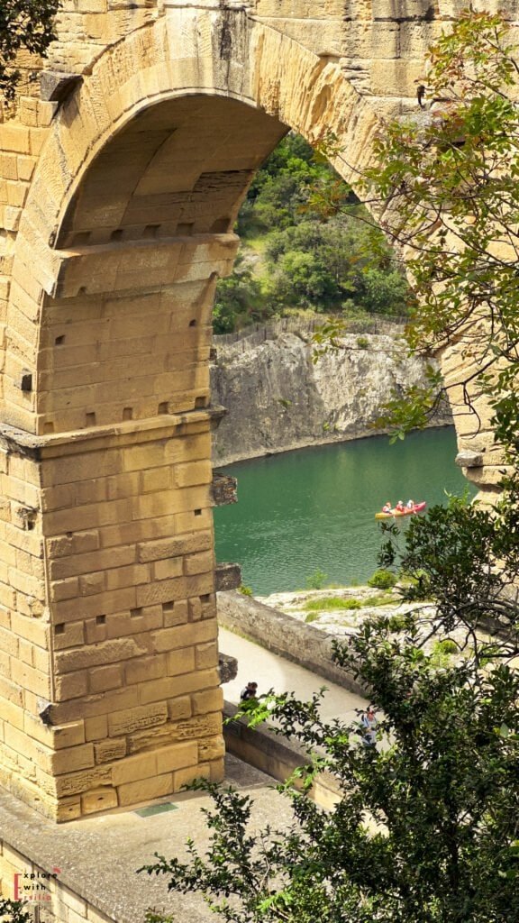 Close-up view of a massive stone arch from the Pont du Gard aqueduct, framing a glimpse of the turquoise Gardon River below. A small group of people in a red and yellow canoe paddles across the calm water, while leafy branches partially obscure the foreground.