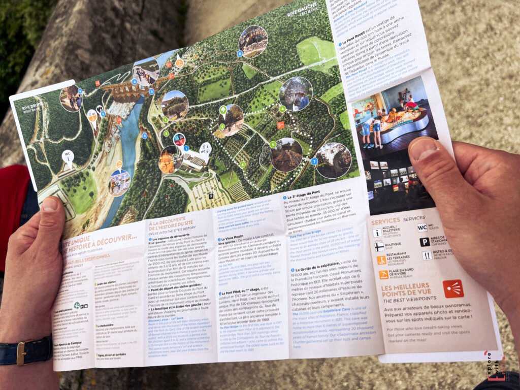 Close-up of a person holding a colorful tourist map of the Pont du Gard site in France, featuring illustrated trails, numbered points of interest, and photos of key attractions. The map includes service icons, French text, and recommended viewpoints, with the surrounding pathway and stone ledge visible in the background.