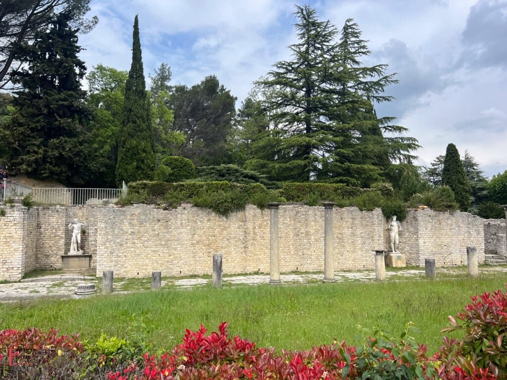 A row of Roman columns and two classical statues stand before a stone wall in the ancient site of Vaison-la-Romaine, France. The lush backdrop includes manicured hedges and tall evergreens, while vibrant red and green foliage decorates the foreground under a partly cloudy sky.